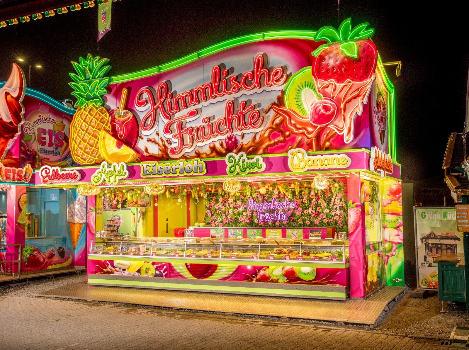 Schaustellerwagen Himmlische Früchte der Familie Eiserloh bei Nacht – pink-grünes Neonschild mit Schokofrüchten, Liebesäpfeln und Fruchtauslage